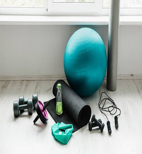 Group of different exercising equipment on white home gym floor. Fitness ball, round foam roller, resistance exercise latex band, jumping rope, dumbbells, yoga mat. Fit lifestyle concept.